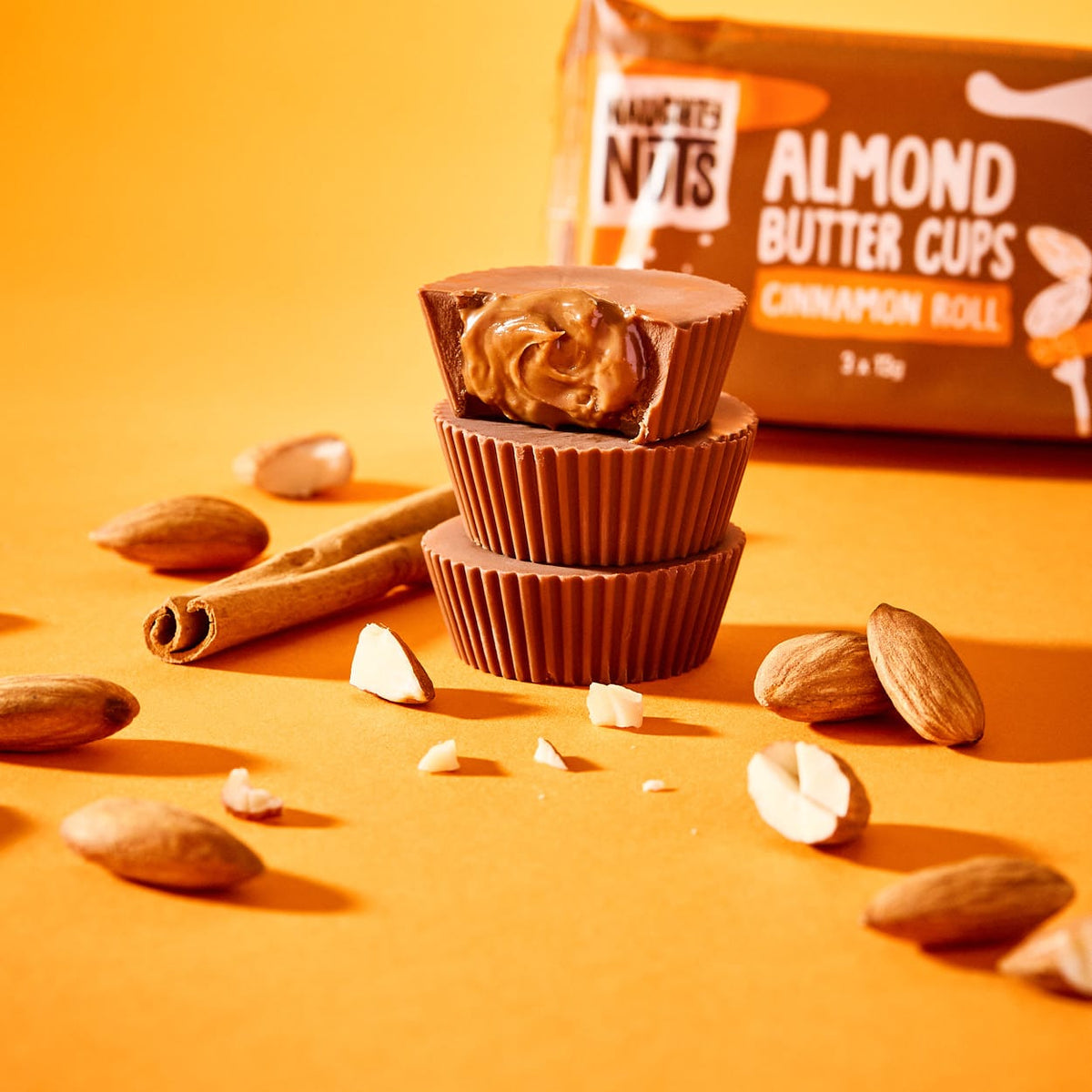 Drei Bio Nutbutter Cups gestapelt, der oberste ist angebissen und zeigt die Füllung, umgeben von Mandeln und Zimtstangen
