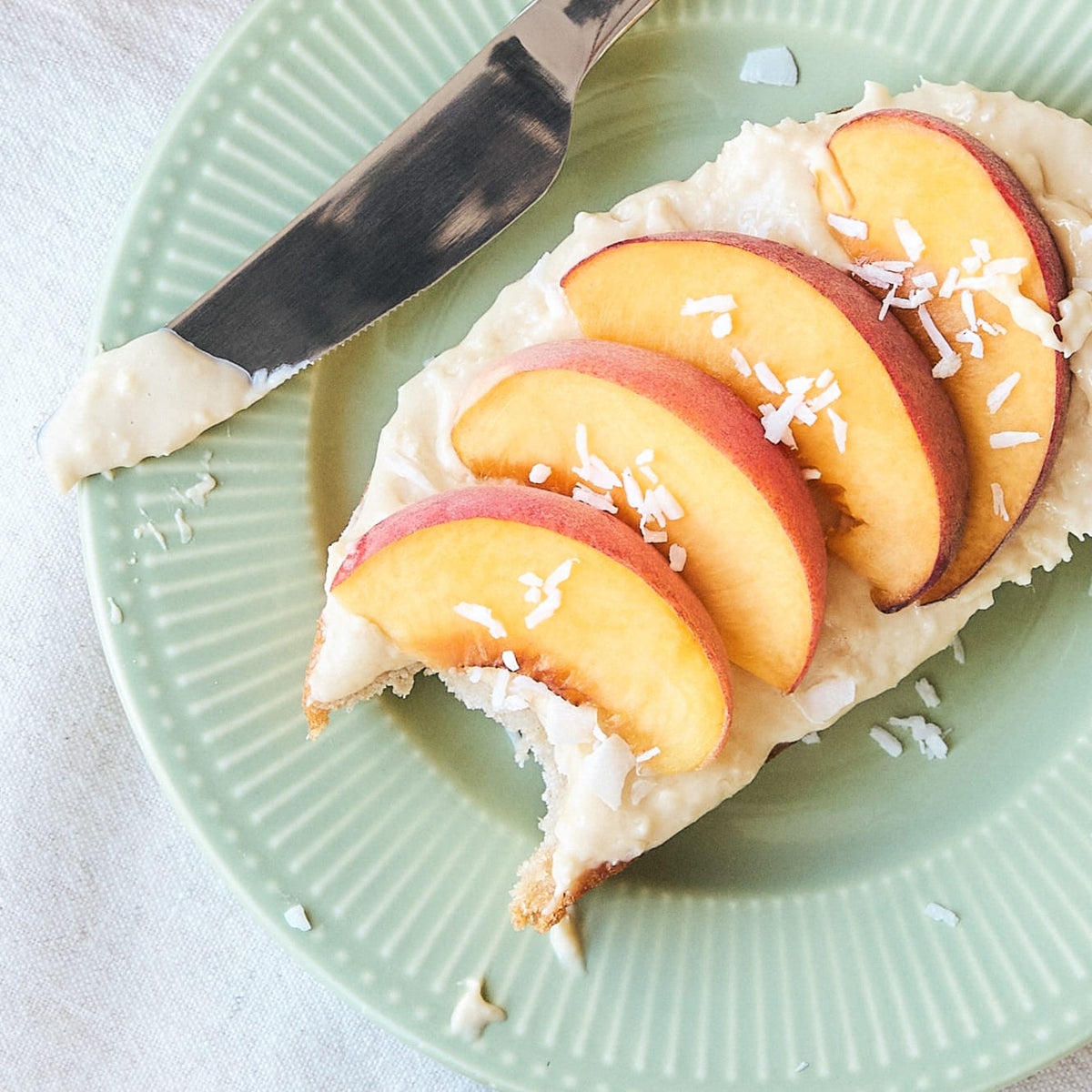 Happy Coconut Creme auf Toast mit Pfirsichscheiben und Kokosraspeln, ein Messer liegt daneben