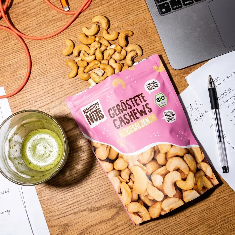 Naughty Nuts geröstete Cashews gesalzen Packung mit verstreuten Cashews, Laptop, Notizen und Getränk